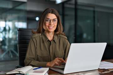 Happy mature middle aged professional business woman investor working on laptop computer in office,...