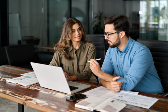 Middle aged female manager teaching young male worker looking at computer discussing ai online business software at office meeting. Two busy colleagues working together talking using laptop in office.