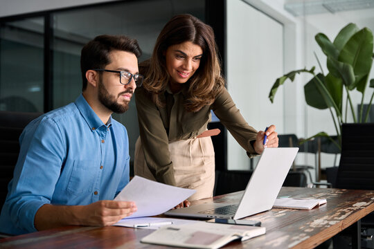 Busy business people using laptop talking in office. Professional mature business woman manager explaining corporate software elearning solutions helping young employee at work. Candid shot.