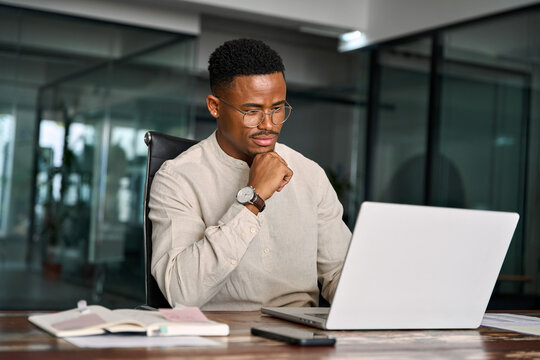 Young male businessman looking at computer thinking on online investment or software technology. Busy professional African business man company employee using laptop working at office.