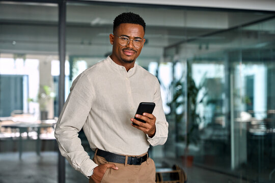 Young Black businessman entrepreneur using smartphone looking at camera with cell phone at work. Smiling busy professional African American business man standing in office holding mobile cellphone.