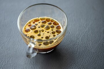 Close-up of a glass cup filled with hot brewed coffee topped with frothy bubbles on a textured dark surface with natural lighting
