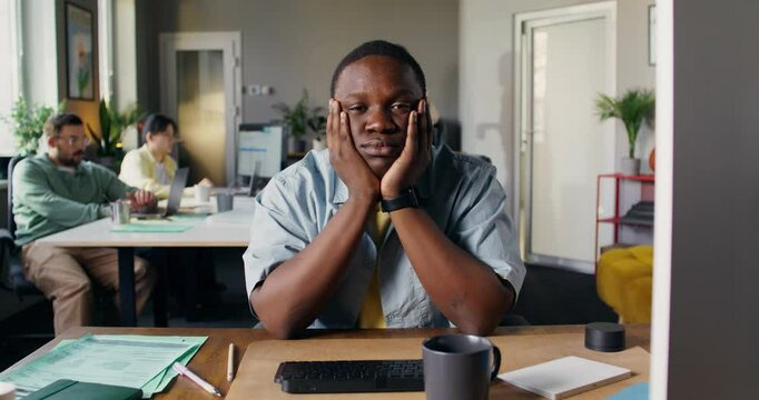 An employee looks at the camera sitting at his desk in the office. His colleagues are moving around the office in a fast-paced motion picture. Hustle and bustle during the working day