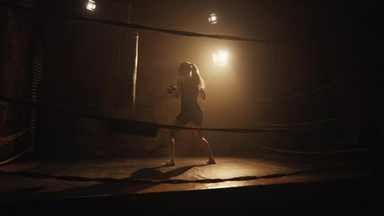 Self-defense and fitness, woman boxing with punch bag in gym, silhouette in dark . Slender blonde sportswoman with ponytail moving active in box ring, training and preparing to fight, cinematic shot - Powered by Adobe
