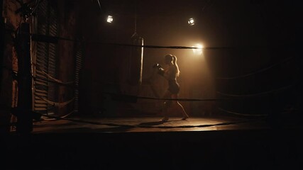 Cinematic slow motion shot with attractive slender woman on boxing ring in dark. Athletic blonde lady punching punch bag and training for stay slender, beautiful and healthy, do professional sport - Powered by Adobe