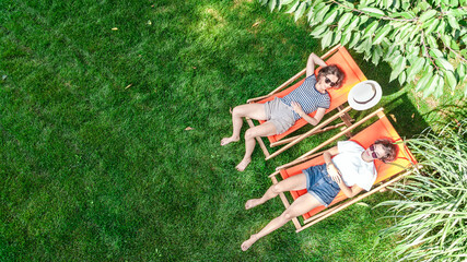 Young girls relax in summer garden in sunbed deckchairs on grass, women friends have fun outdoors in green park on weekend, aerial drone view from above