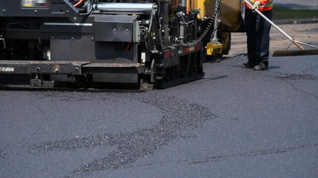 Asphalt paving machine laying fresh blacktop on a road. A worker in a high-visibility vest is using a rake or asphalt lute to smooth and guide the material, ensuring even coverage