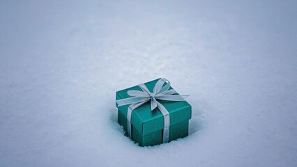 Green box with a silver ribbon sits in the snow. The box is wrapped in a shiny silver ribbon and is sitting on top of the snow
