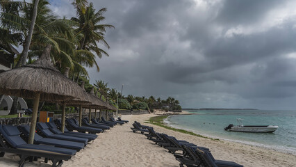 Rows of deck chairs with mattresses under straw umbrellas on a sandy beach. A boat in the turquoise...