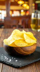 A close-up of a wooden bowl filled with crispy potato chips on a dark slate serving board in a lively pub setting