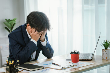 A frustrated young Asian businessman sits at his office desk using a laptop, showing signs of stress, exhaustion, and worry. © Worawi