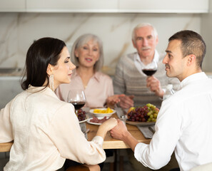 Happy young couple looking into each other's eyes sitting at table while senior parents looking at them gladly in the kitchen