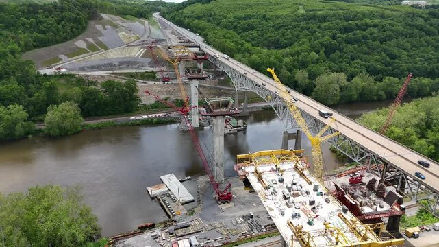 Aerial view of highway I-80 in Pennsylvania showing a bridge over a river surrounded by lush forests and mountains, with heavy traffic and massive construction of a second highway bridge nearby.