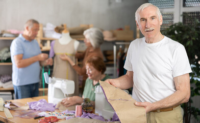 In training tailoring workshop for older adults, elderly man smiling while showing paper pattern...