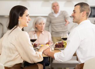 Couple sharing close moment at dining table, holding hands and locking eyes, while perplexed parents watching in background at family home