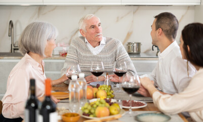 Smiling elderly man engaging in lively conversation with wife, grown son and daughter-in-law at family table with wine and fruits in cozy home kitchen