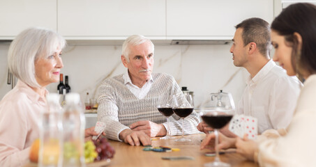 Joyful senior parents and young couple sitting around table playing with cards and drinking wine in goblets at home