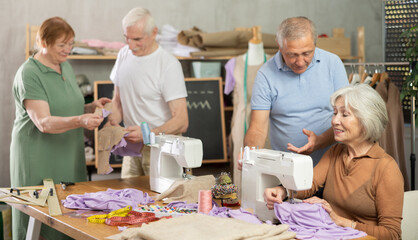 Obraz premium Elderly men and women spending time together sewing in cozy workroom, friendly chatting while using machines, cutting fabric and paper templates during hands-on leisure time