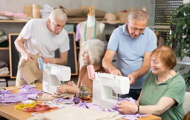 Elderly men and women spending time together sewing in cozy workroom, friendly chatting while using machines, cutting fabric and paper templates during hands-on leisure time