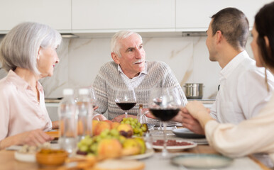 Mature couple are sitting at table with younger spouses friend, drinking wine and enjoying snacks, chatting merrily, raise glass to, celebrating important event.