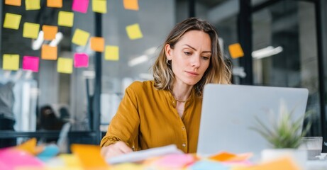 Focused businesswoman working on a laptop in a colorful office environment with sticky notes on the glass wall. Deadline and meeting concept.