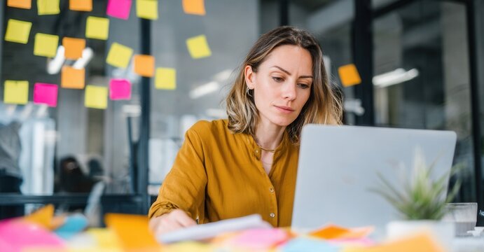 Focused businesswoman working on a laptop in a colorful office environment with sticky notes on the glass wall. Deadline and meeting concept.