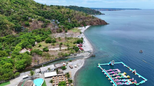 Aerial 4K morning drone footage of Mabayuan Beach in Olongapo, Zambales, Philippines, showing turquoise waves lapping white sand with rugged mountains and a distant city skyline.