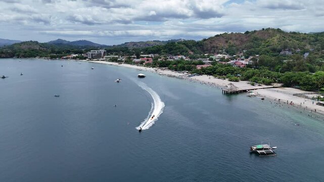 Aerial 4K morning drone footage of Mabayuan Beach in Olongapo, Zambales, Philippines, showing turquoise waves lapping white sand with rugged mountains and a distant city skyline.