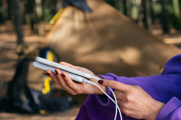 Close-up woman with smartphone and portable power bank in hands, against the background of a tent...