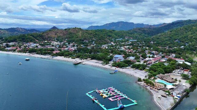 Aerial 4K morning drone footage of Mabayuan Beach in Olongapo, Zambales, Philippines, showing turquoise waves lapping white sand with rugged mountains and a distant city skyline.