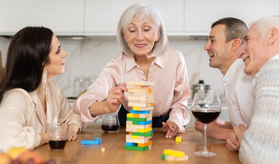 Group of mature spouses and middle-aged male visitor with girlfriend is playing board game. Senior married couple build tower with wooden blocks bricks with younger spouses