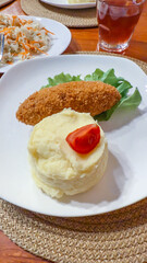 Mashed potatoes topped with cherry tomato half, breaded cutlet on lettuce leaf, with a glass of red juice and side of coleslaw salad vertical