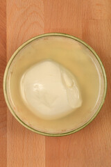 A single, round ball of fresh mozzarella cheese floats in a clear liquid within a bowl on a wooden surface