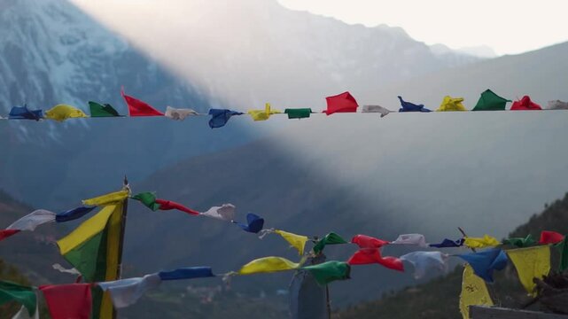 Slow motion shot of colorful Buddhist Prayer flags waving in the wind in front of snowy Himalayan mountain peaks with sun rays during the sunset as seen from Tayul monastery in Keylong, Lahaul Valley.