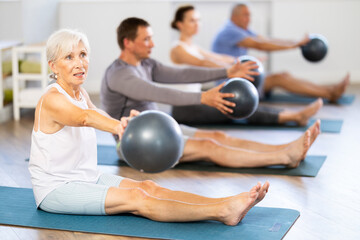 Group of elderly people in sportswear exercising Pilates with soft balls while sitting on mats in fitness studio