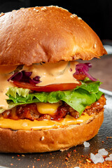 A close-up of a gourmet burger with a sesame seed bun, melted cheese, lettuce, tomato, pickles, red cabbage, and creamy sauce. The burger is placed on a dark surface with coarse salt and spices 