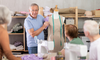 Smiling experienced male teacher in sixties leading sewing course for seniors, attaching fabric to...