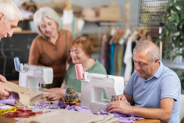 Enthusiastic elderly man participating in sewing class for seniors, stitching fabric on machine, assembling garment pieces while fellow learners cutting patterns and fitting garments on mannequin..