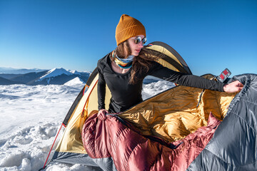 Young woman takes out a sleeping bag from a tent in the snowy mountains. Winter camping scene
