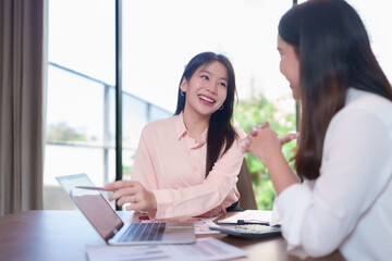 Fototapeta premium Asian Women Discussing Business Strategy on Laptop with Smiling Faces in Modern Office Brainstorming Ideas and Reviewing Data for Financial Growth