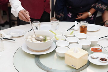 A restaurant waiter is pouring soup with fish balls into each bowl on a round table for restaurant guests