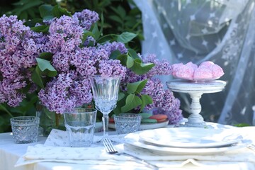 Beautiful lilac flowers, sweets, plates, cutlery and glasses on table in garden