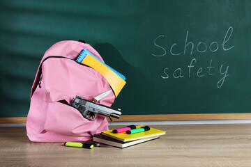 Backpack with gun, knife and stationery on wooden table near blackboard. School safety