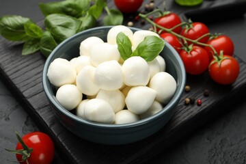 Tasty mozzarella cheese balls in bowl, basil, peppercorns and tomatoes on black table, closeup