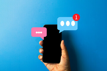 Hand holding a smartphone with chat speech bubbles and a new message notification icon.,Concept of mobile communication, social media, texting, online conversation, and app alerts.,Blue background.