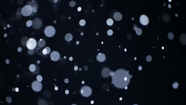 Soft snow and particles glide in the air slowly start falling in the dark, black background.