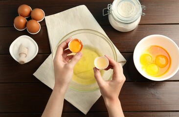 Woman separating egg yolk from white over glass bowl at wooden table, top view
