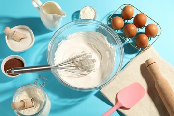 Whisk with whipped cream, eggs and other products on light blue table, flat lay