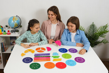 Two kids and a female teacher sit at a table exploring math concepts using brightly colored...