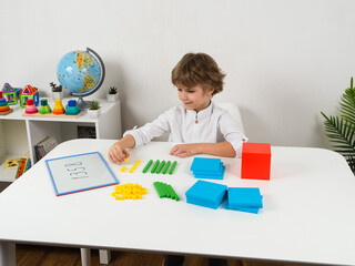A young boy with a cheerful expression is sitting at a white table, learning about place value and counting using colorful base ten blocks. He is surrounded by the manipulatives 